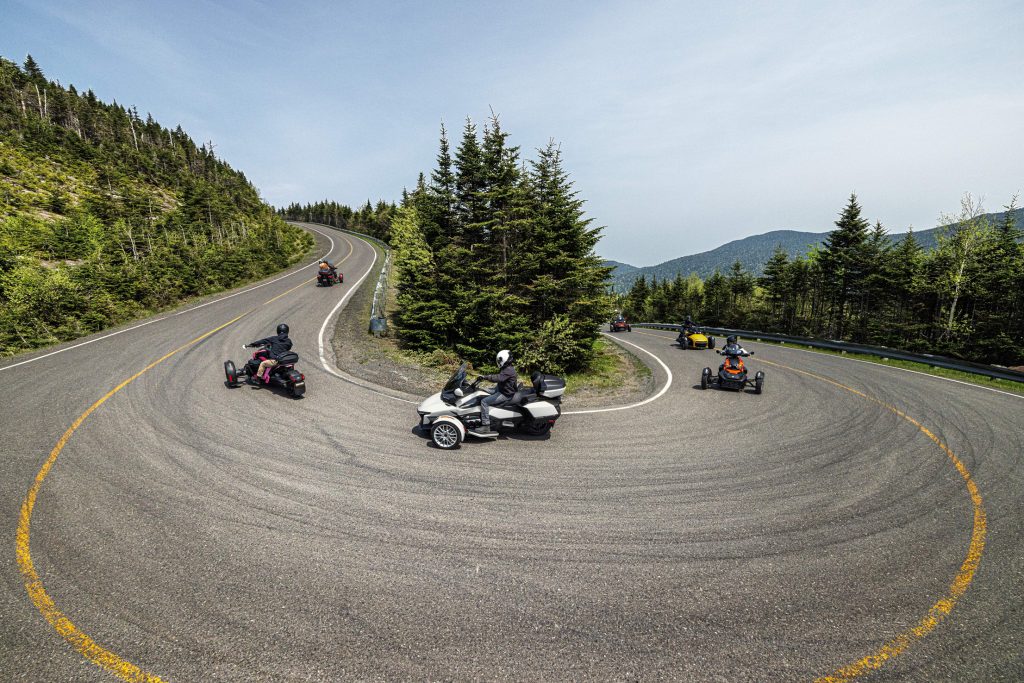 Plusieurs modèles de Can-Am Spyder et Ryker en mouvement dans un virage serré en montagne, illustrant la stabilité et le plaisir de conduite sur route sinueuse.