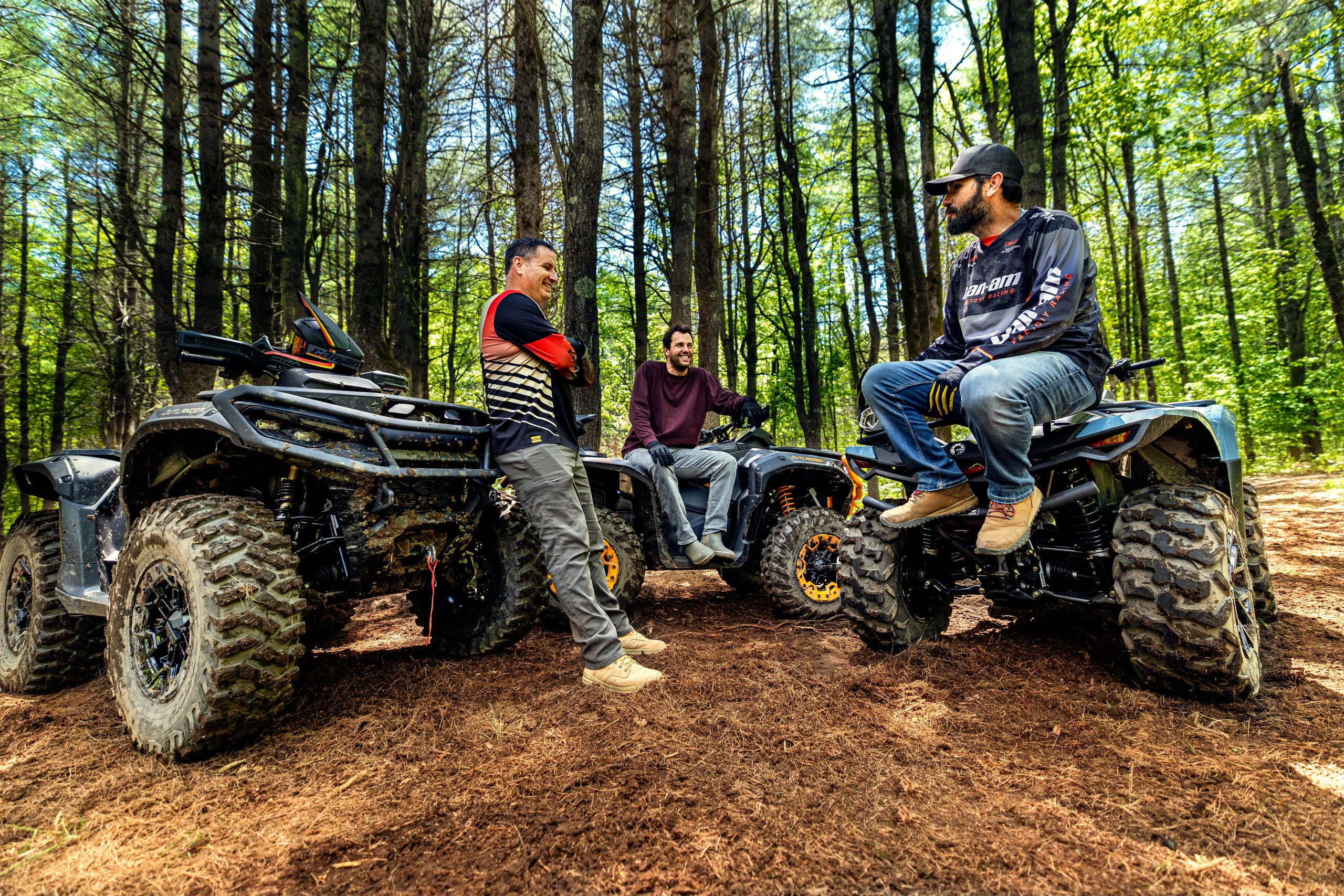 Trois hommes discutent en forêt à côté de leurs VTT Can-Am Outlander boueux lors d'une randonnée tout-terrain.